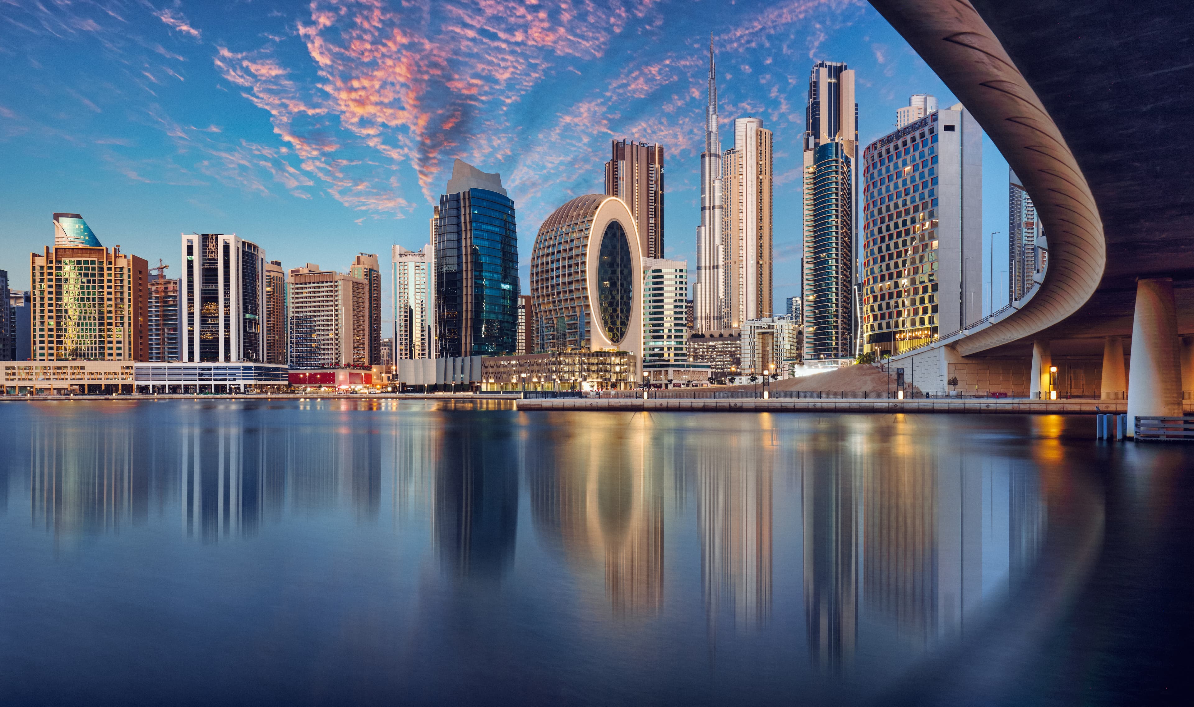 A serene view of Dubai's skyline reflected in calm waters, highlighting its futuristic architecture and distinctive circular structures under a beautifully painted sky.