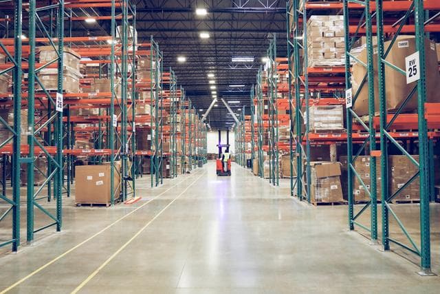 The image shows a large warehouse with tall shelving units stocked with pallets and boxes. A forklift is positioned in the centre aisle, which is wide and well-lit, allowing for easy movement and efficient storage operations. The orderly setup indicates a well-organised logistics or distribution facility.