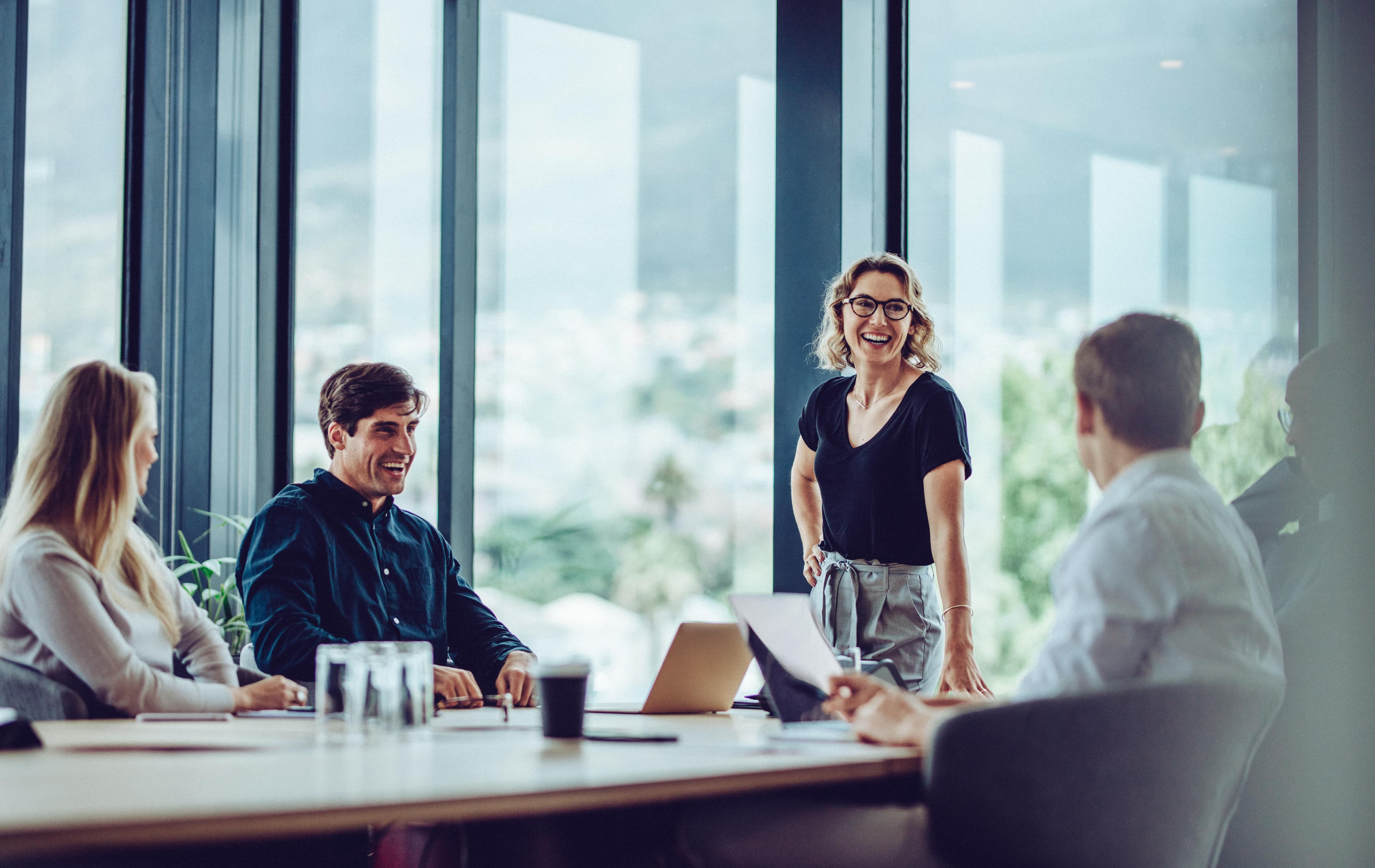 A team meets in a modern office, with one person standing and leading the discussion, creating a collaborative and positive atmosphere. Large windows provide natural light.