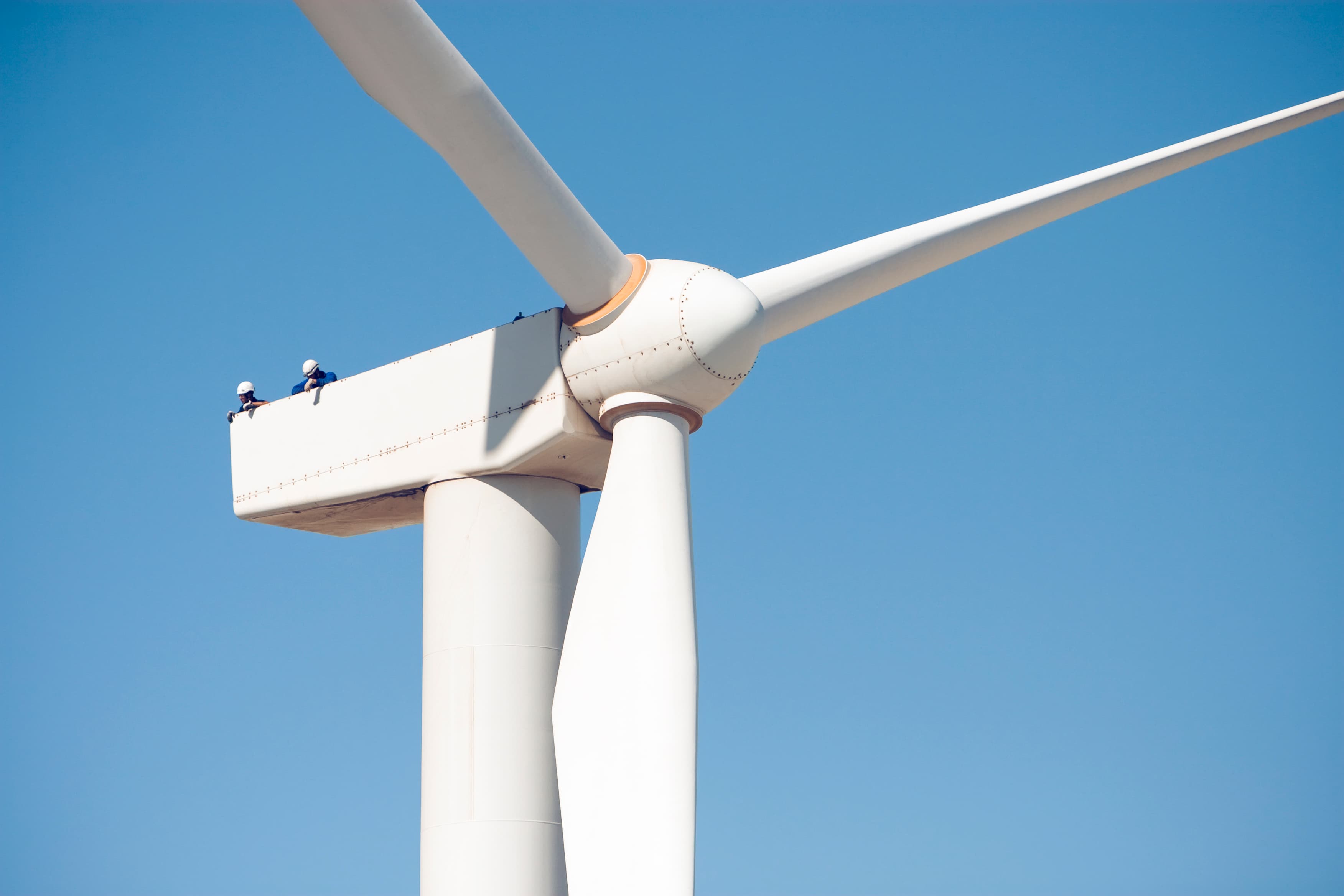 This image shows two workers atop a large wind turbine, performing maintenance or inspection. The turbine’s blades are prominently visible against a clear blue sky, highlighting the scale of wind energy infrastructure and the importance of safety and technical expertise in the wind industry.