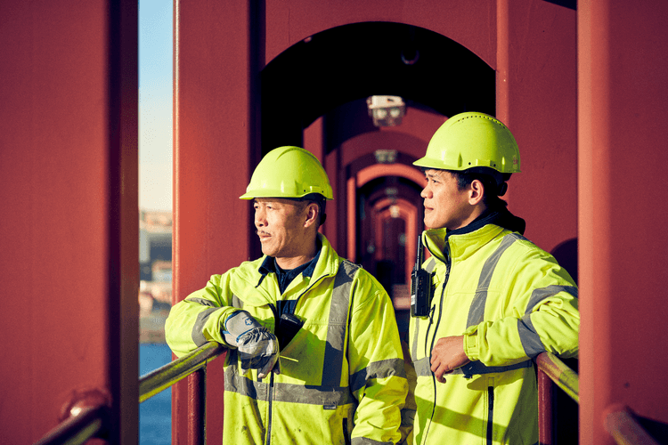 Two workers in high-visibility jackets and helmets stand on a metal walkway, looking thoughtfully into the distance. The image highlights teamwork, safety, and focus within an industrial or maritime setting.
