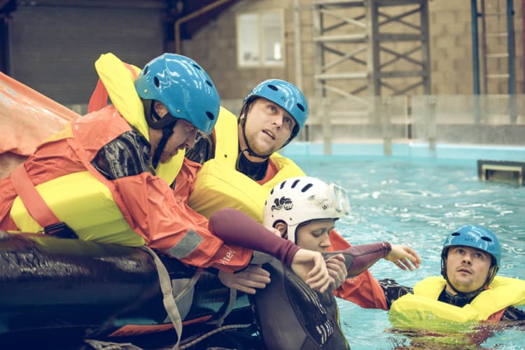 a sea survival training exercise, with participants in safety helmets and life jackets practising life raft drills in a controlled water environment.